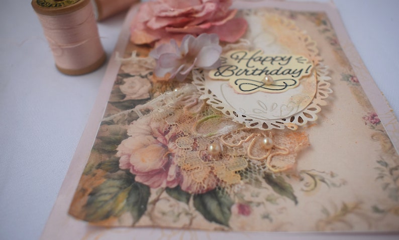 Card is flat on white background. Base is pink shimmer under vintage rose pattern. Left side has handmade & prima flowers in pinks and hand-dyed lace snippet.  Vintage oval frame is in middle showing vintage effect. Sentiment is Happy Birthday.