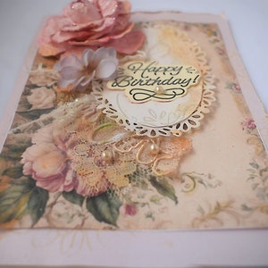 Card is flat on white background. Base is pink shimmer under vintage rose pattern. Left side has handmade & prima flowers in pinks and hand-dyed lace snippet.  Vintage oval frame is in middle showing vintage effect. Sentiment is Happy Birthday.