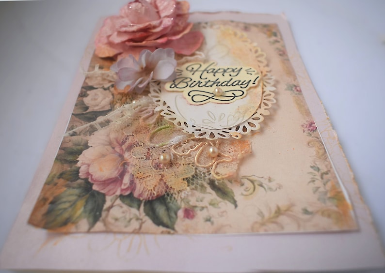 Card is flat on white background. Base is pink shimmer under vintage rose pattern. Left side has handmade & prima flowers in pinks and hand-dyed lace snippet.  Vintage oval frame is in middle showing vintage effect. Sentiment is Happy Birthday.