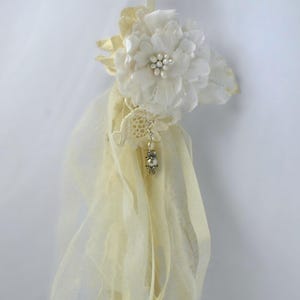 May include: A white fabric flower with pearl accents hangs from a long, cream-colored fabric streamer. The streamer is made of multiple layers of sheer fabric.
