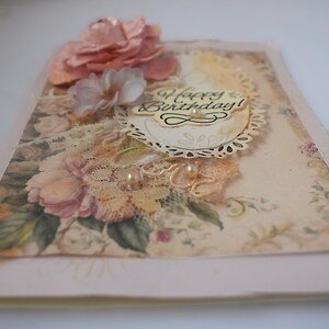 Card is flat on white background. Base is pink shimmer under vintage rose pattern. Left side has handmade & prima flowers in pinks and hand-dyed lace snippet.  Vintage oval frame is in middle showing vintage effect. Sentiment is Happy Birthday.