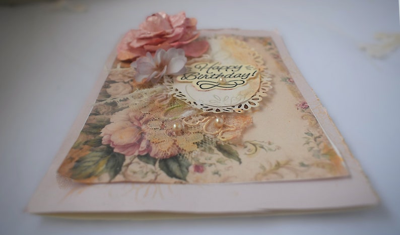 Card is flat on white background. Base is pink shimmer under vintage rose pattern. Left side has handmade & prima flowers in pinks and hand-dyed lace snippet.  Vintage oval frame is in middle showing vintage effect. Sentiment is Happy Birthday.