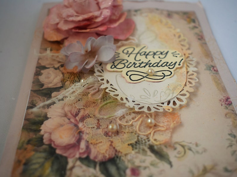 Card is flat on white background. Base is pink shimmer under vintage rose pattern. Left side has handmade & prima flowers in pinks and hand-dyed lace snippet.  Vintage oval frame is in middle showing vintage effect. Sentiment is Happy Birthday.