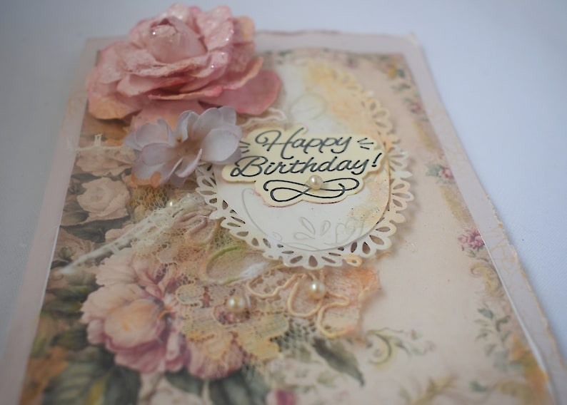 Card is flat on white background. Base is pink shimmer under vintage rose pattern. Left side has handmade & prima flowers in pinks and hand-dyed lace snippet.  Vintage oval frame is in middle showing vintage effect. Sentiment is Happy Birthday.