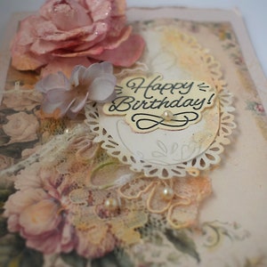 Card is flat on white background. Base is pink shimmer under vintage rose pattern. Left side has handmade & prima flowers in pinks and hand-dyed lace snippet.  Vintage oval frame is in middle showing vintage effect. Sentiment is Happy Birthday.