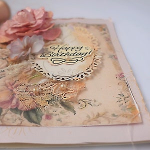 Card is flat on white background. Base is pink shimmer under vintage rose pattern. Left side has handmade & prima flowers in pinks and hand-dyed lace snippet.  Vintage oval frame is in middle showing vintage effect. Sentiment is Happy Birthday.