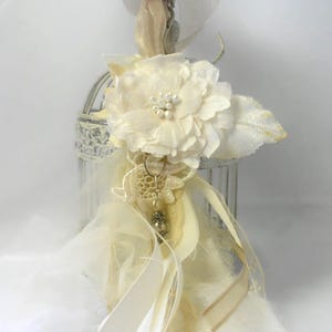 May include: A white birdcage with a white fabric flower, lace, and ribbon accents. The birdcage is decorated with a pearl and a small metal charm.