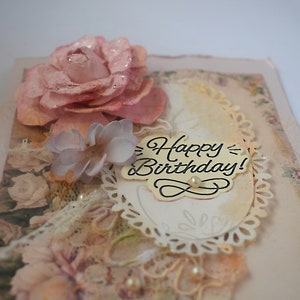 Card is flat on white background. Base is pink shimmer under vintage rose pattern. Left side has handmade & prima flowers in pinks and hand-dyed lace snippet.  Vintage oval frame is in middle showing vintage effect. Sentiment is Happy Birthday.