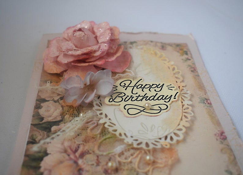 Card is flat on white background. Base is pink shimmer under vintage rose pattern. Left side has handmade & prima flowers in pinks and hand-dyed lace snippet.  Vintage oval frame is in middle showing vintage effect. Sentiment is Happy Birthday.