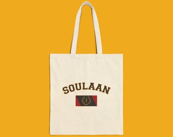 Könnte beinhalten: Eine cremefarbene Stofftasche mit dem Wort SOULAAN in dunkelbrauner Schrift im College-Stil. Unter dem Text befindet sich eine rechteckige Grafik mit schwarzem und rotem Hintergrund sowie einem goldenen Emblem eines Schwertes und eines Lorbeerkranzes.