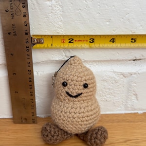 May include: A tan crochet peanut-shaped plush toy with dark brown feet, black bead eyes, and a stitched black smile. A metal keychain attachment is at the top. A yellow measuring tape shows the toy is approximately 2.5 inches wide.