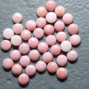 May include: A collection of round, polished gemstones in varying shades of pink and white. The cabochon stones are scattered across a gray surface, showcasing their smooth, domed surfaces. The gemstones are likely for jewelry making.