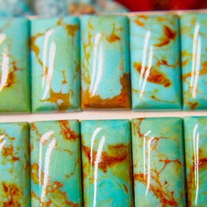 May include: Close-up of rectangular turquoise gemstones with a polished, glossy finish. The stones have a vibrant blue-green hue with brown veining, arranged in two rows. The gemstones are likely for jewelry making.