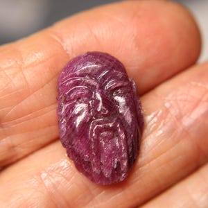 Op de afbeelding: Een gesneden, dieppaarse edelsteen in de vorm van een menselijk gezicht. Het gezicht heeft gedetailleerde kenmerken, waaronder een baard en duidelijke gezichtslijnen. De edelsteen wordt in een hand gehouden.