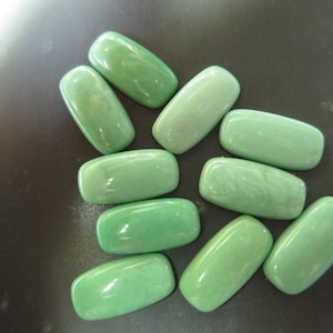 May include: A collection of eleven polished, light green rectangular stones. The stones have rounded edges and a smooth, glossy surface. The arrangement is scattered on a dark surface, with varying shades of green.