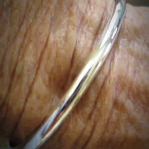 Op de afbeelding: Een zilveren armband om de pols van een persoon. De armband is glad en glanzend.