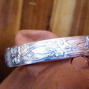 May include: A silver bracelet with an intricate floral design. The bracelet is made of polished metal and has a smooth, rounded surface.
