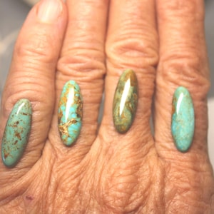 May include: Four oval-shaped turquoise gemstones with brown and gold matrix patterns. The stones are displayed on a hand, showcasing their polished surfaces and natural variations. These gemstones are suitable for jewelry making.