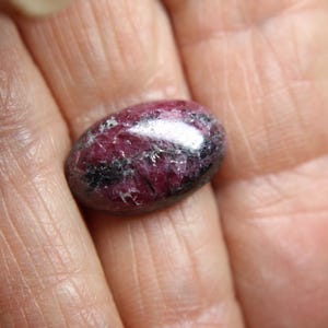 May include: A polished, oval-shaped gemstone with a deep burgundy hue, accented by streaks of black and a reflective, silvery sheen. The stone's surface displays a natural, textured pattern, suggesting a mineral specimen.