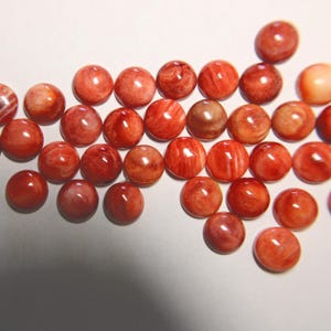 May include: A collection of round, polished gemstones in shades of red and orange. The stones have a smooth, glossy surface and vary in color intensity, with some showing internal patterns. The gemstones are arranged on a white surface.