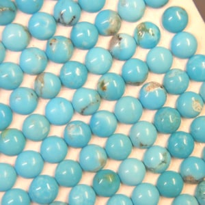 May include: A collection of numerous round, smooth turquoise cabochons arranged in a grid pattern. Each stone features a vibrant sky-blue color with unique, subtle dark brown or grey matrix veining patterns visible on the polished, domed surfaces.