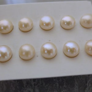 May include: A close-up of ten round, light peach-colored pearls arranged in two rows on a white card. Each pearl has a subtle luster and is set on a gold-colored base. The pearls are the focal point of the image.