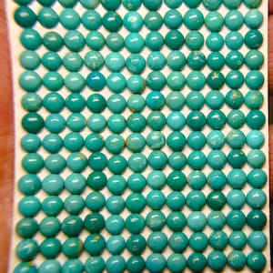 May include: A collection of small, round turquoise gemstones. The stones vary in shades of blue and green, with some showing natural veining. The gemstones are arranged in a grid pattern, suggesting they are ready for use in jewelry or crafts.