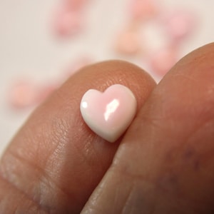 May include: A small, heart-shaped bead with a glossy finish. The bead is primarily white with a soft pink center, held between two fingers. Other similar beads are slightly out of focus in the background.