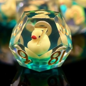 May include: A clear, multi-sided die with a yellow rubber ducky inside. The die has gold-colored numbers and a teal and yellow background. The number 7 is visible on the front of the die. Other dice are visible in the background.