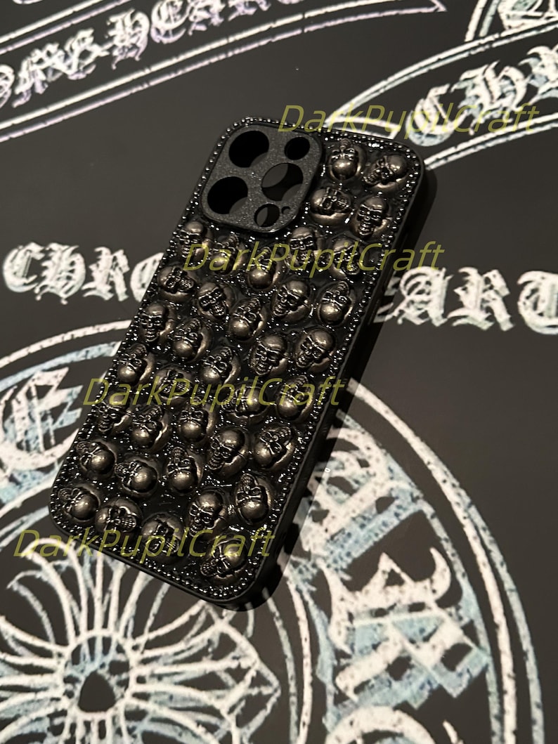 May include: A black phone case with a skull design. The case is covered in small, silver skulls. The case has a cutout for the camera lens. The text 'DarkPupilCraft' is printed on the case.