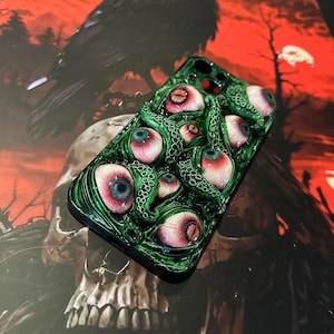 Op de afbeelding: Groene telefoonhoes met meerdere realistische oogballen met rode, blauwe en witte details. De hoes heeft een textuur, tentakelachtig ontwerp. De achtergrond bevat een raaf en een schedel, wat een griezelig thema cre&euml;ert.