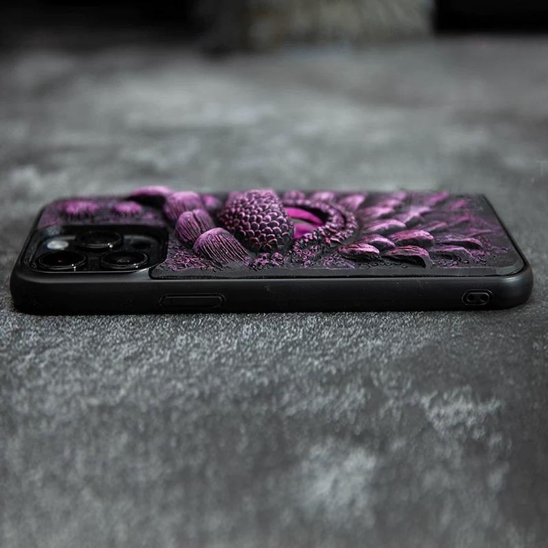May include: A black smartphone case featuring a three-dimensional, textured purple dragon eye design with intricate scale patterns. The case includes a cutout for the rear camera array and covers the back and sides of the device.