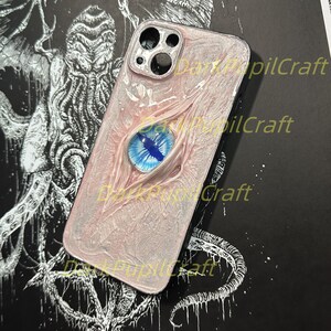 May include: A white and pink phone case with a blue eye design. The case has a black camera lens cutout and the text "Dark Pupil Craft" is visible on the case.