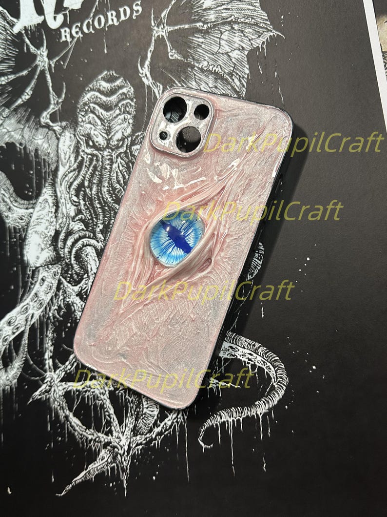 May include: A white and pink phone case with a blue eye design. The case has a black camera lens cutout and the text "Dark Pupil Craft" is visible on the case.