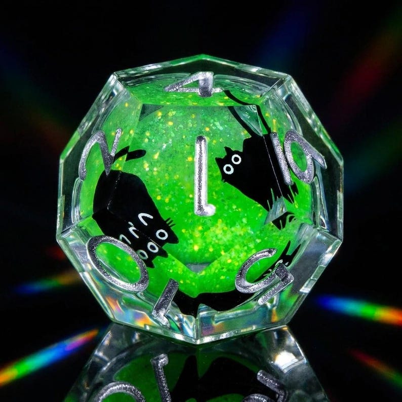 May include: A clear, multi-faceted D20 die with a vibrant green interior filled with glitter. Black cat silhouettes are featured inside the die. The numbers are silver. The die is reflected on a black surface.