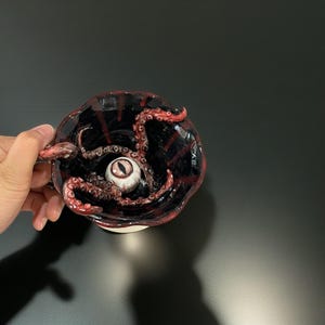 以下が含まれることがあります： 黒と赤のセラミック製のマグカップで、中に白い目と赤い触手が付いています。マグカップはカップの形で、持ち手があります。