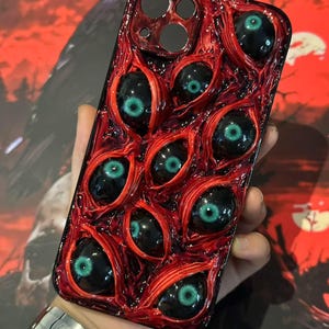 May include: A red and black phone case featuring a design of multiple eyeballs with green irises. The case has a glossy finish and a textured appearance, creating a unique and eye-catching aesthetic. The case is held in a person's hand.