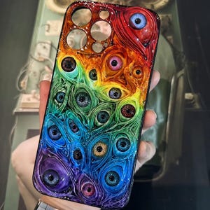 May include: A rainbow-colored phone case featuring a pattern of multiple eyes. The case transitions from red to purple, with each eye having a unique iris color. The case has cutouts for the camera and buttons.