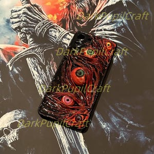 Puede incluir: Una funda de tel&eacute;fono negra con un dise&ntilde;o 3D de m&uacute;ltiples ojos rojos con pupilas verdes. La funda est&aacute; sobre un fondo de una ilustraci&oacute;n de fantas&iacute;a oscura de un esqueleto sosteniendo una espada.