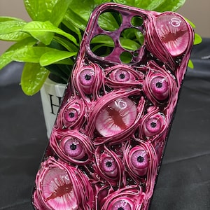 Puede incluir: Una funda de teléfono rosa y negra con un diseño de muchos ojos. Los ojos son 3D y tienen un acabado brillante.