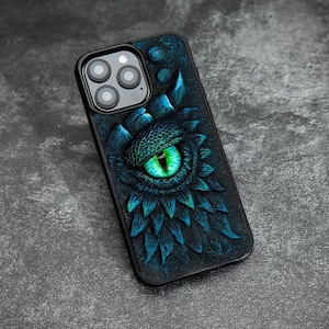 May include: A black smartphone case featuring a three-dimensional, textured design of a large, glowing green dragon eye surrounded by dark blue, scale-like patterns. The case includes a cutout for a triple-lens camera system on the back.