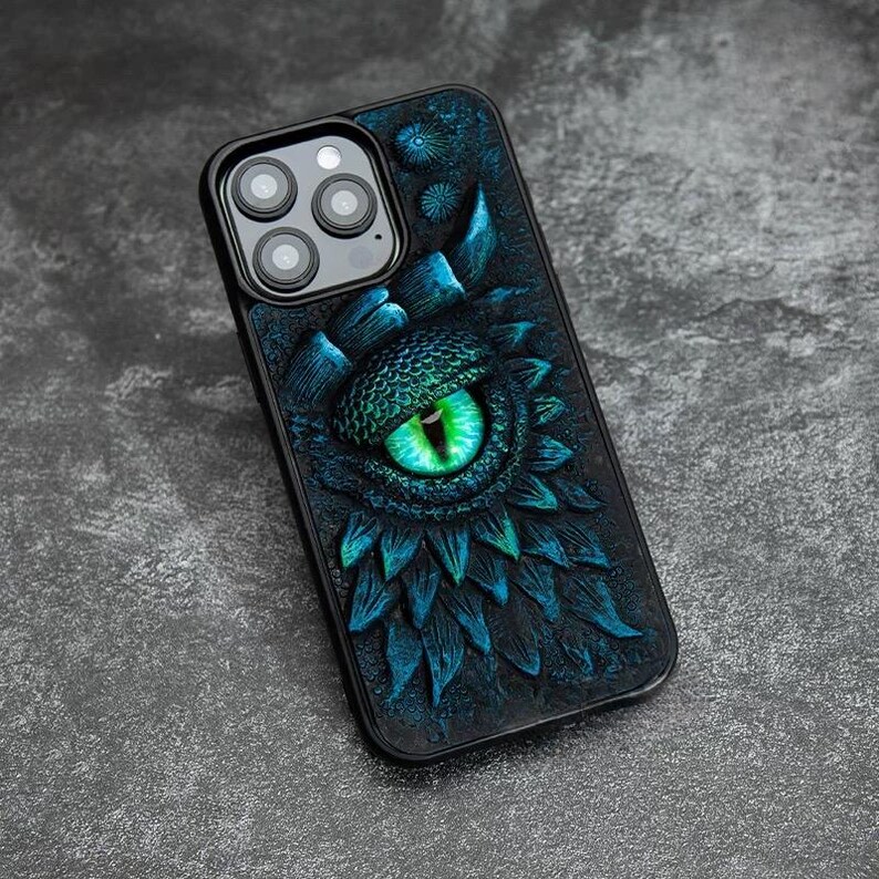 May include: A black smartphone case featuring a three-dimensional, textured design of a large, glowing green dragon eye surrounded by dark blue, scale-like patterns. The case includes a cutout for a triple-lens camera system on the back.