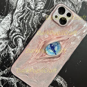 May include: A pink and white phone case with a blue eye and a black and white illustration of a creature on the back. The case has a camera lens cutout and the text "DarkPupilCraft" is visible.