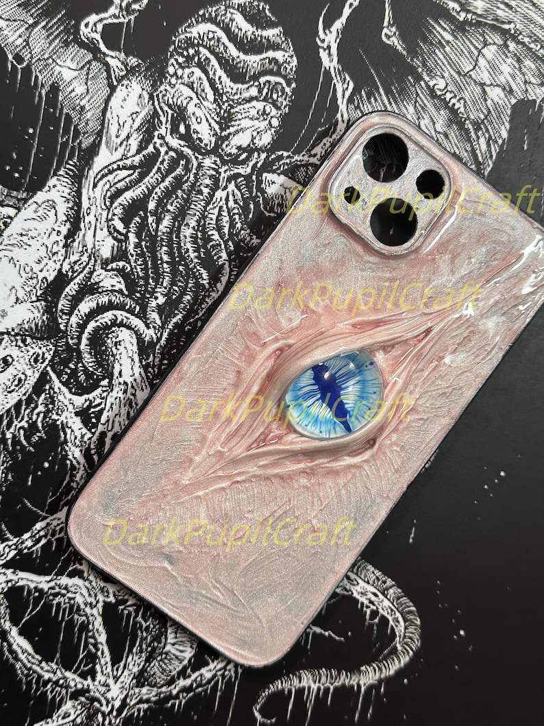May include: A pink and white phone case with a blue eye and a black and white illustration of a creature on the back. The case has a camera lens cutout and the text "DarkPupilCraft" is visible.