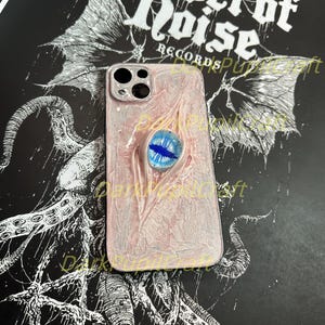 May include: A white and pink iPhone case with a blue eye design. The case has a textured surface and a black border. The case is on a black background with a white design.
