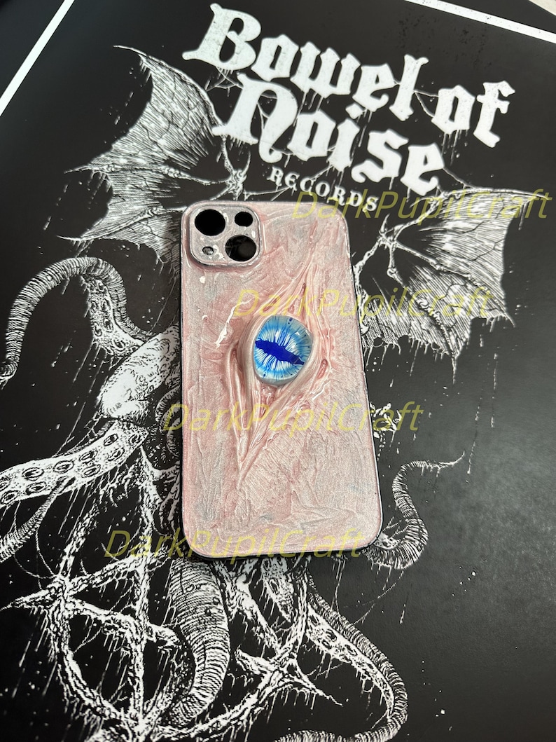 May include: A white and pink iPhone case with a blue eye design. The case has a textured surface and a black border. The case is on a black background with a white design.