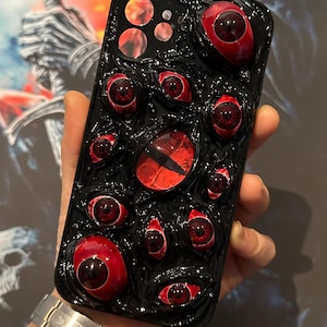 May include: A black phone case with a 3D design of red eyes. The eyes are raised and have a glossy finish.