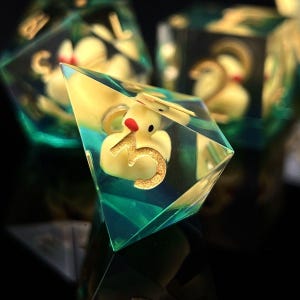May include: A set of handmade dice featuring a yellow rubber ducky with a red beak and gold number five inside a clear, iridescent resin. The dice have a teal and yellow color scheme and are set against a dark background.