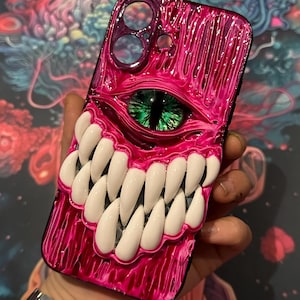 Peut inclure: Une coque de téléphone rose et noire avec un design sculpté en 3D d'un œil et de dents de monstre. L'œil est vert et les dents sont blanches.