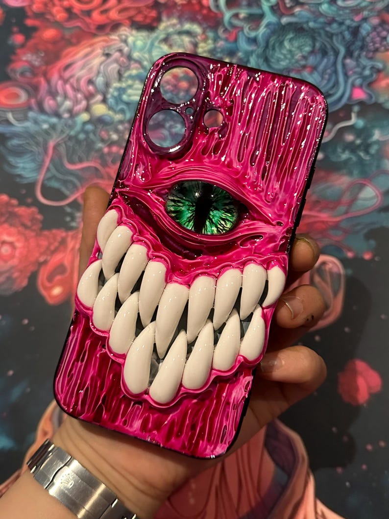 May include: A pink and black phone case with a 3D sculpted design of a monster's eye and teeth. The eye is green and the teeth are white.